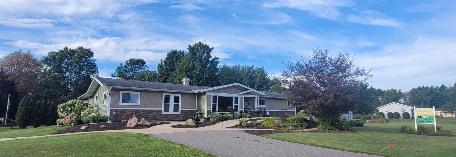 Single-story Evergreen Park assisted living building with a landscaped front lawn, paved driveway and entrance ramp under a partly cloudy sky.