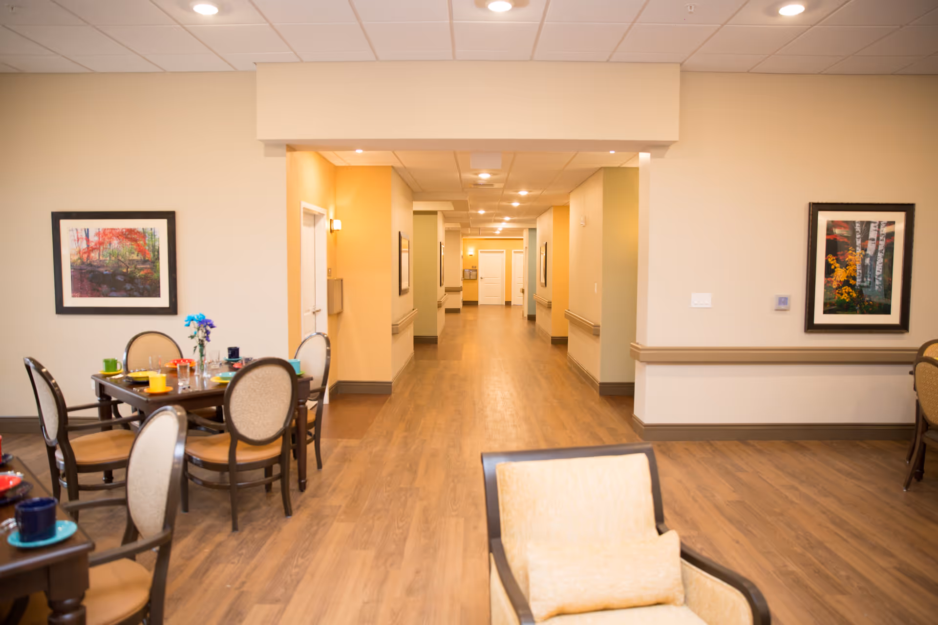 Interior view of a senior living facility hallway with wooden flooring and beige walls. On the left side, there are dining tables set with colorful cups and plates, and chairs with cushioned seats. The hallway is well-lit with ceiling lights and has framed artwork on the walls.