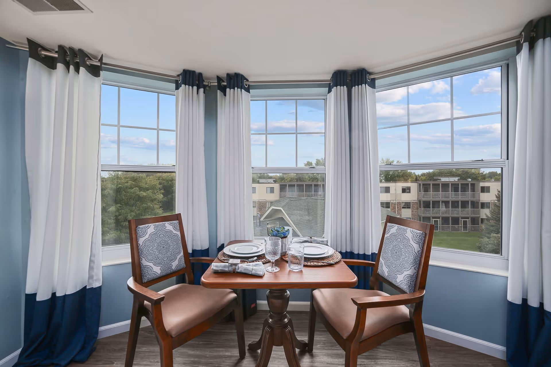 A small dining area with a wooden table set for two, featuring two wooden chairs with patterned cushions. The area is surrounded by three large windows with white and navy blue curtains, offering a view of trees and nearby buildings under a partly cloudy sky.