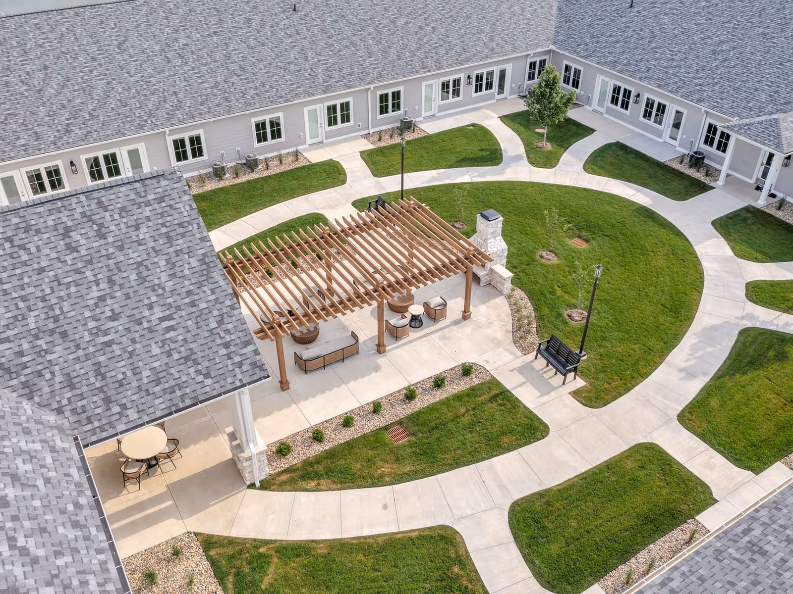 Aerial view of a landscaped courtyard with winding walkways, a wooden pergola seating area, benches, and the surrounding senior living building.