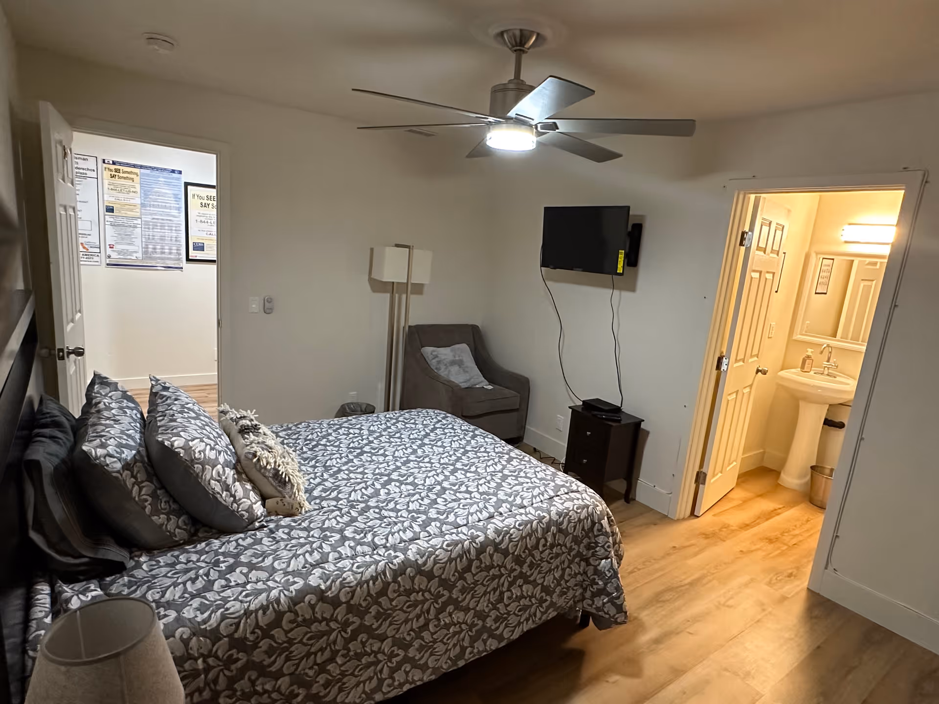 A bedroom with a bed covered in a patterned gray and white bedspread and multiple pillows. There is a ceiling fan with a light above the bed. A gray armchair is placed in the corner next to a small dark wooden nightstand with a flat-screen TV mounted on the wall above it. The room has light-colored wooden flooring. An open door reveals a bathroom with a pedestal sink, mirror, and bright lighting. Another open door shows a hallway with posters on the wall.