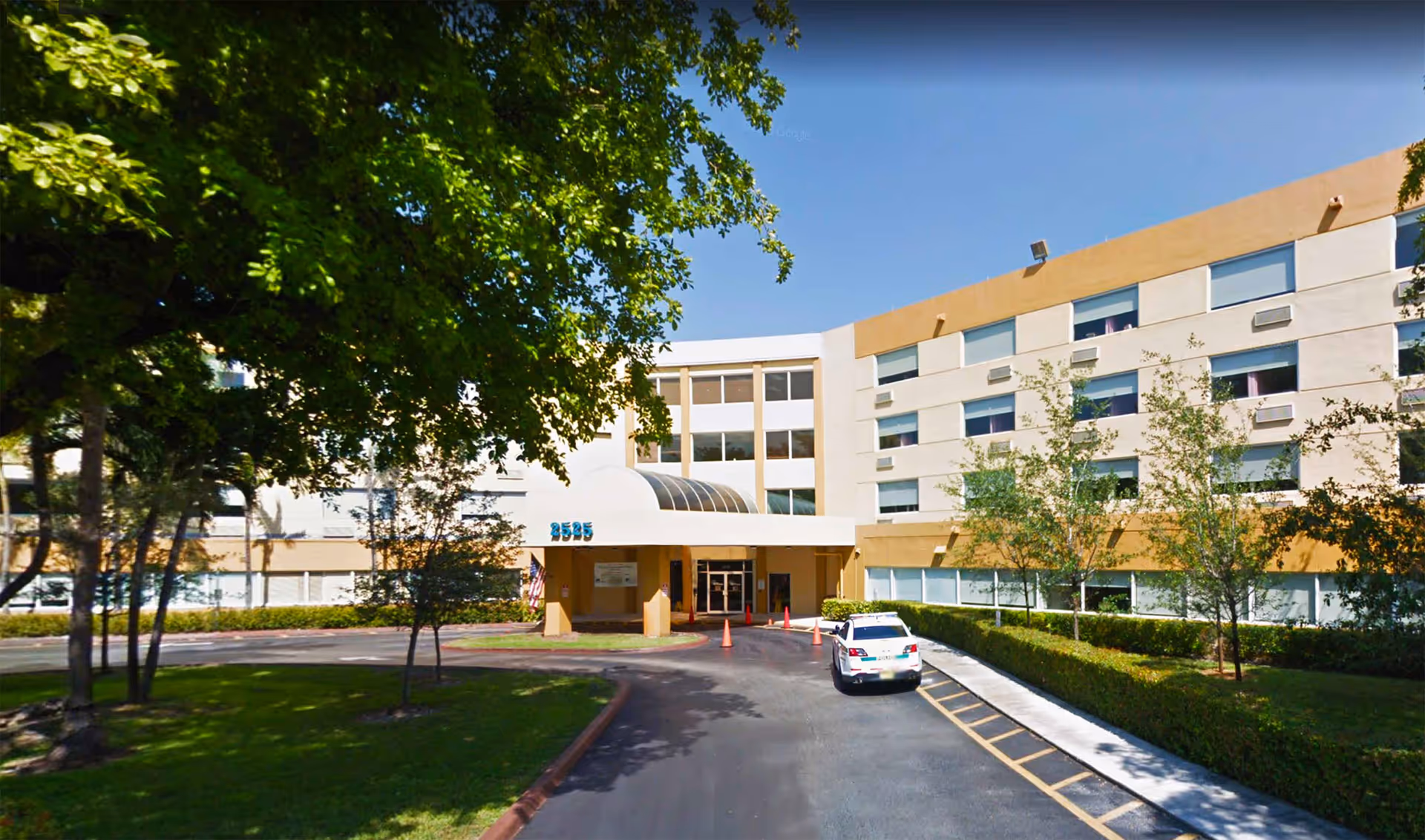 Front exterior view of West Gables Health Care Center showing a multi-story building with beige and white walls, several windows, a covered entrance, a driveway with a parked white car, and surrounding trees and greenery under a clear blue sky.