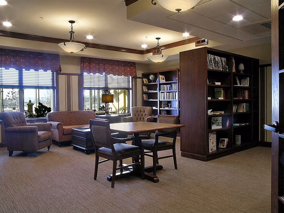 A cozy common area with a table and four chairs in the center, surrounded by upholstered armchairs and a sofa near windows with blinds and valances. There are bookshelves filled with books and decorative items along the walls. The room is softly lit by ceiling lights and has a carpeted floor.