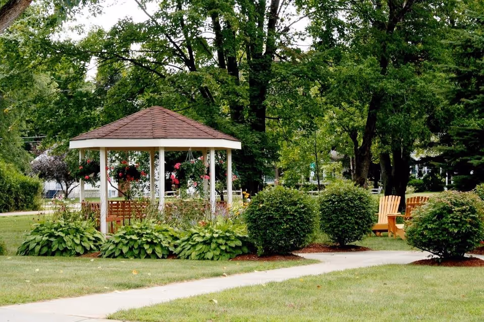 A peaceful outdoor garden area with a white gazebo featuring a brown roof, surrounded by green bushes and plants. There are hanging flower baskets inside the gazebo and wooden chairs placed on the grass nearby. Tall trees provide shade in the background.