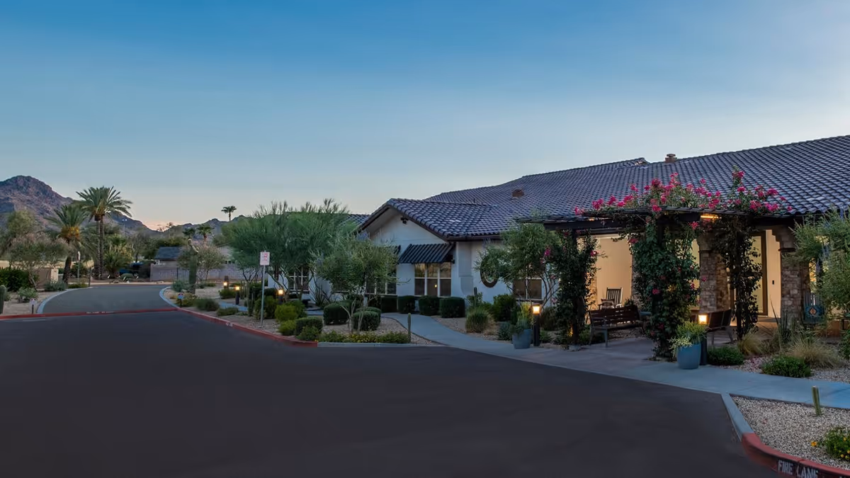 Exterior view of a single-story building with a tiled roof at dusk, surrounded by landscaped greenery including palm trees and bushes. A covered seating area with climbing plants and benches is visible near the entrance. Mountains can be seen in the background under a clear sky.