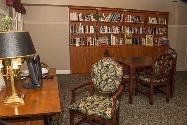 A cozy interior room with a wooden table and chairs upholstered in floral fabric. A large wooden bookshelf filled with books is against the wall. A wooden desk with a lamp and a computer monitor is positioned near a window with blinds.
