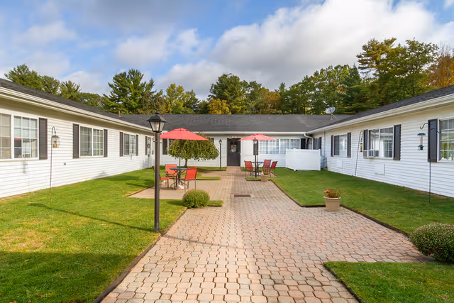 Sunlit courtyard lined by single-story white buildings with a central paved walkway, red umbrella patio tables, lawn, and trees.