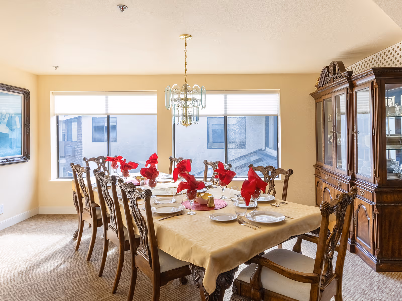 A formal dining room with a long wooden table covered with a beige tablecloth. The table is set with white plates, silverware, glasses with red napkins folded inside, and a centerpiece. There are eight ornate wooden chairs around the table. Two large windows with blinds allow natural light into the room. A wooden china cabinet filled with dishes is against the right wall, and a framed picture hangs on the left wall. A chandelier hangs above the table.