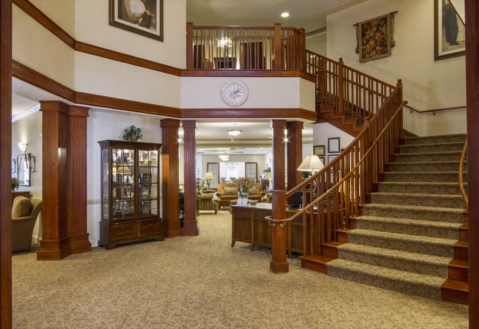 Interior view of a senior living facility lobby with a carpeted staircase on the right, wooden columns, a glass display cabinet, and a seating area with sofas and lamps in the background. The walls are decorated with framed artwork and a clock above the entrance to the seating area.