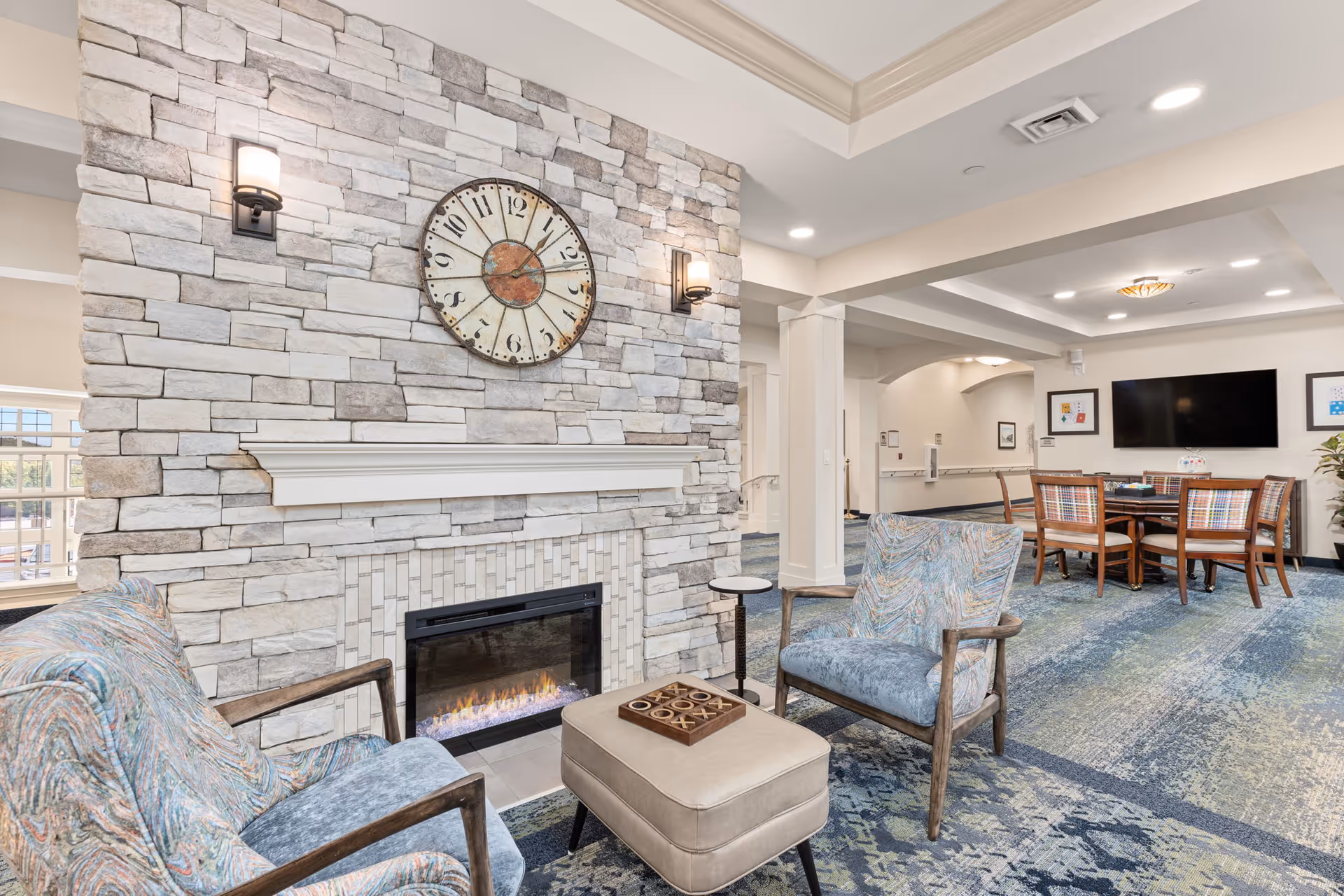A cozy senior living facility common area with a stone fireplace, a large wall clock above it, two patterned armchairs, a beige ottoman with a tic-tac-toe game on top, and a round table with four chairs in the background. The room has a blue and green patterned carpet and a flat-screen TV mounted on the wall.