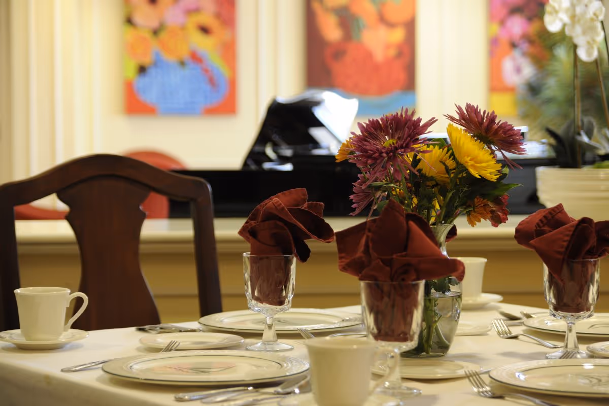 A dining table set with plates, cups, silverware, and glasses with folded maroon napkins. A vase with colorful flowers is placed in the center of the table. In the background, there is a black grand piano and colorful floral paintings on the wall.