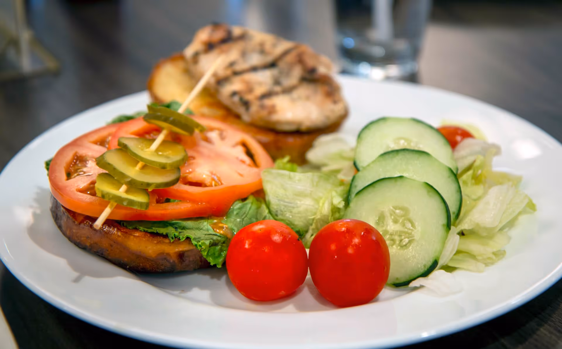 A plate with a sandwich featuring grilled chicken, lettuce, tomato slices, and pickles on a bun, accompanied by a side salad with cucumber slices, cherry tomatoes, and lettuce.