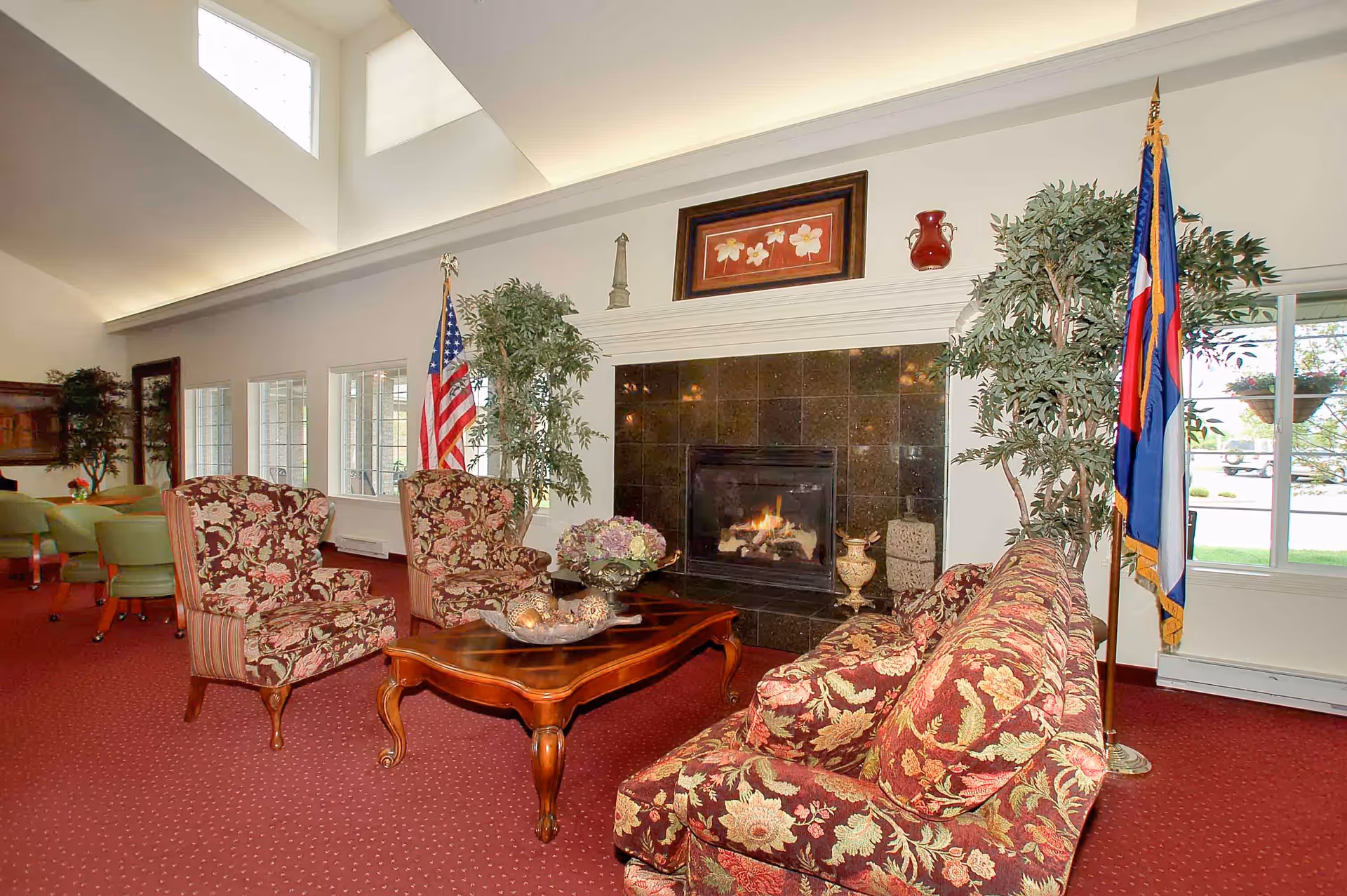 A cozy living room area with floral patterned armchairs and a matching sofa arranged around a wooden coffee table. A fireplace with a black tile surround is lit, and above it hangs a framed floral artwork. Two flags stand on either side of the fireplace, and large windows let in natural light. The room has a red carpet and high ceilings with skylights.