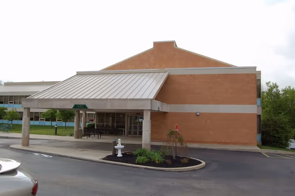 Entrance of a brick building with a covered porte-cochère, small landscaped island with a fountain, and a parking area.