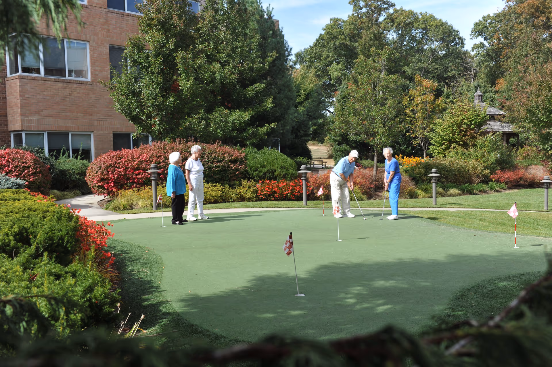 Four elderly people playing golf on a putting green surrounded by lush greenery and colorful bushes, with a brick building visible in the background under a clear sky.