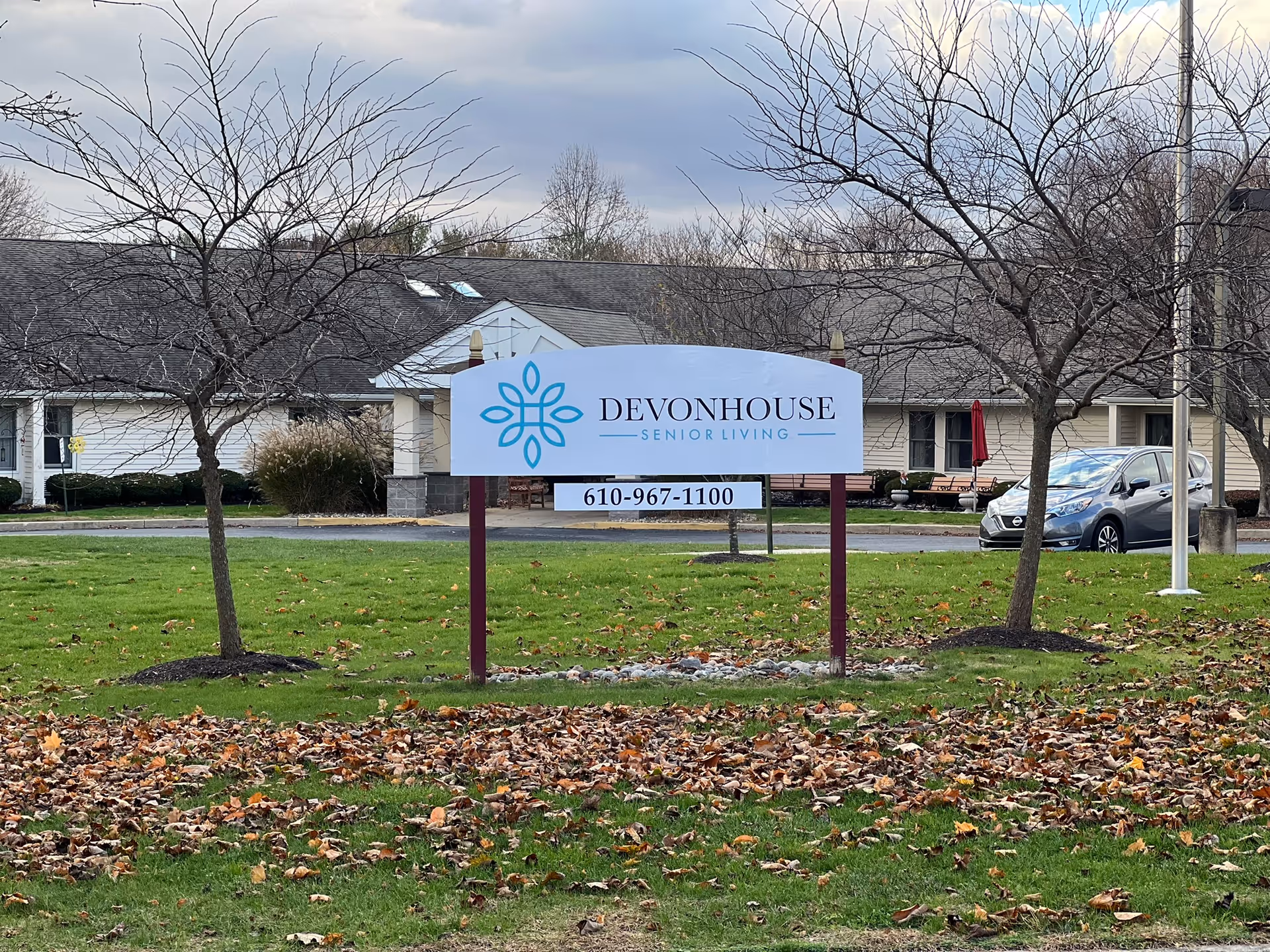 Outdoor view of a sign for DevonHouse Senior Living with a phone number 610-967-1100, set on a grassy area with fallen leaves and leafless trees, with a building and parked car in the background under a cloudy sky.