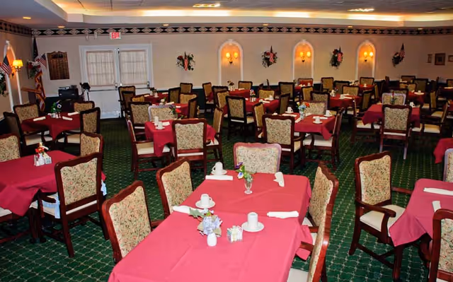 A dining room with multiple tables covered in red tablecloths, each set with white cups, napkins, and small flower arrangements. The chairs have floral upholstery. The room has green carpet with a patterned design, wall sconces with lights, and decorative wall hangings.