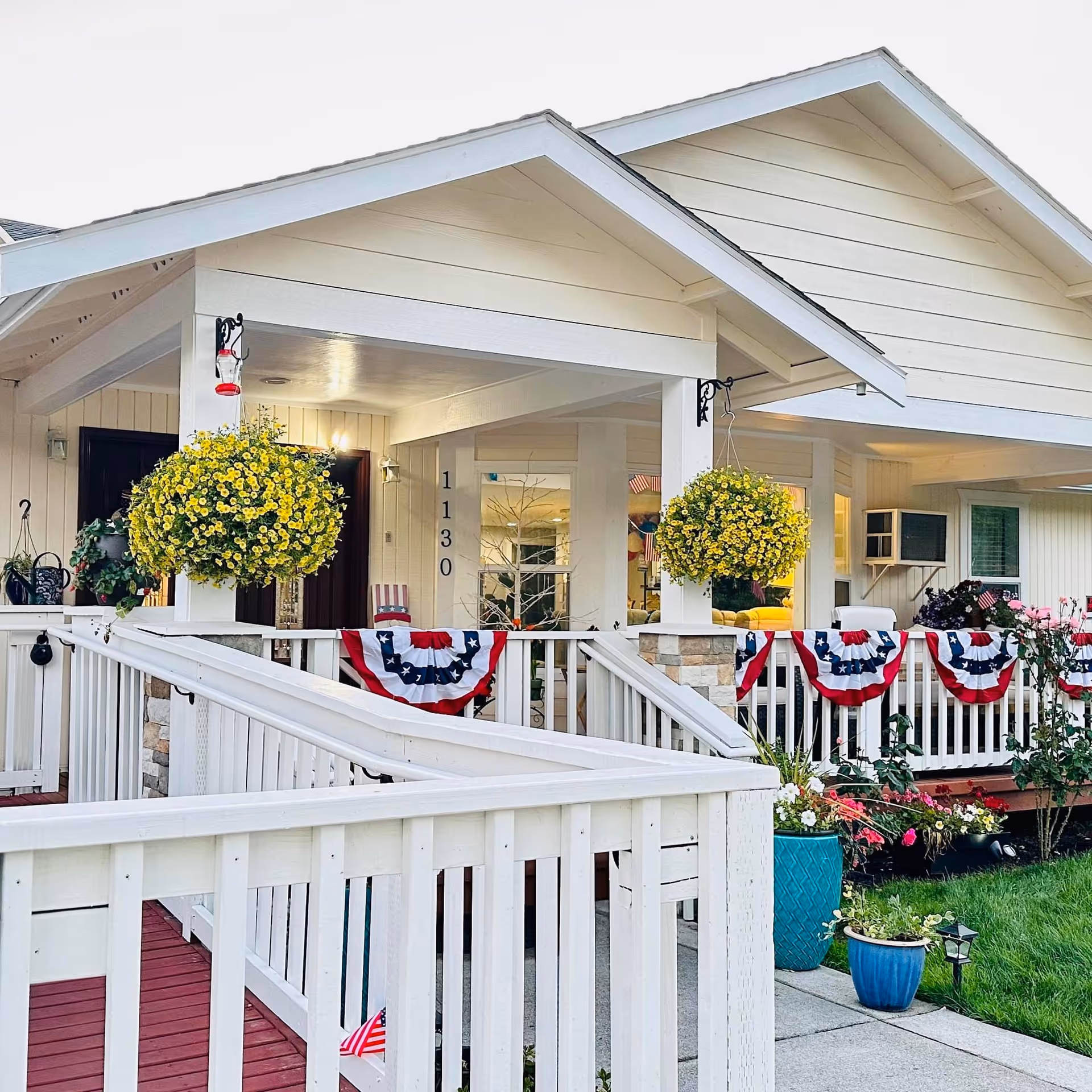 Front exterior view of a white adult family home with a covered porch decorated with patriotic red, white, and blue bunting. Yellow hanging flower baskets and various potted plants adorn the porch and surrounding area. A wheelchair-accessible ramp leads up to the entrance, and the house number 1130 is visible on a pillar.