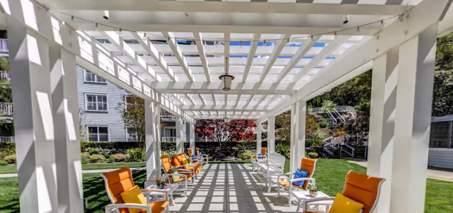 Outdoor seating area under a white pergola with orange cushioned chairs and small tables on a paved pathway, surrounded by green grass and trees with a building visible in the background.