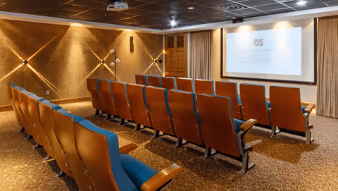 Small movie-style screening room with rows of upholstered theater seats facing a projection screen in a carpeted interior.