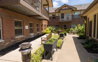 Outdoor courtyard area of a senior living facility with brick buildings on either side, balconies, benches, planters with flowers, and a paved walkway under a clear blue sky.