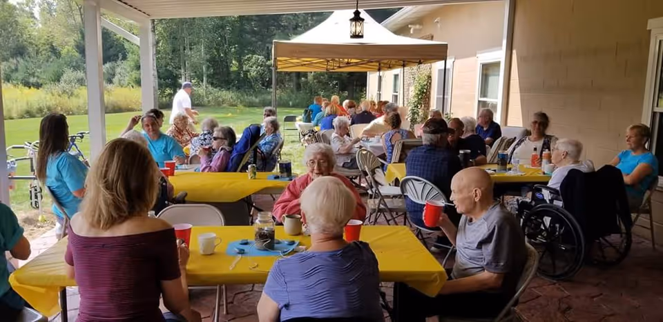 Several elderly residents and caregivers seated at tables with yellow tablecloths under a covered patio, sharing an outdoor meal or gathering.