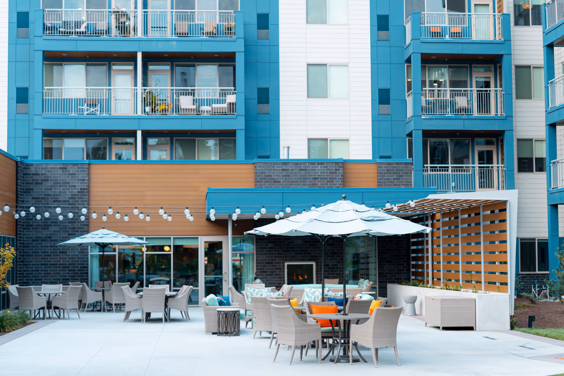 Outdoor patio area of a senior living facility with tables, chairs, umbrellas, string lights, and a fireplace, surrounded by a multi-story building with balconies.