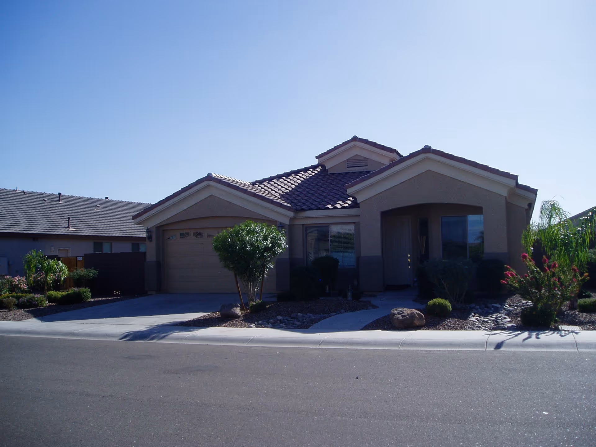 Single-story residential building with a tiled roof, a garage on the left, a front door with a small porch, and landscaped front yard with bushes and rocks under a clear blue sky.