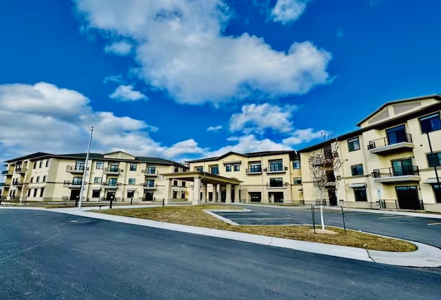 Wide exterior view of a large, modern three-story senior living facility building under a partly cloudy blue sky. The building has multiple balconies and a covered entrance with a driveway and parking area in front.