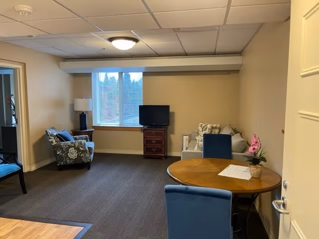A cozy living room area with a round wooden table and blue chairs in the foreground. In the background, there is a window with blinds, a small TV on a wooden stand, a floral armchair with a lamp beside it, and a light-colored sofa with cushions. The room has beige walls and a carpeted floor.
