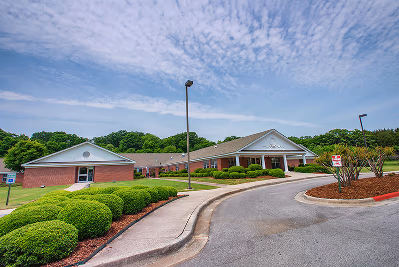 Exterior view of a single-story brick building with a gabled roof, surrounded by neatly trimmed bushes and a curved driveway. The sky is partly cloudy and there are street lamps along the driveway.