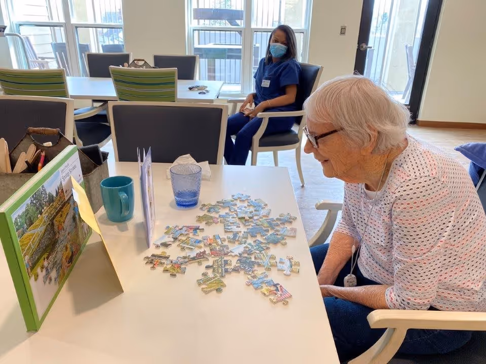 An elderly woman with white hair and glasses is sitting at a table working on a jigsaw puzzle. She is smiling and wearing a white patterned shirt. On the table are puzzle pieces, a blue cup, a glass, and a puzzle box. In the background, a woman in blue scrubs and a face mask is seated on a chair near large windows letting in natural light.