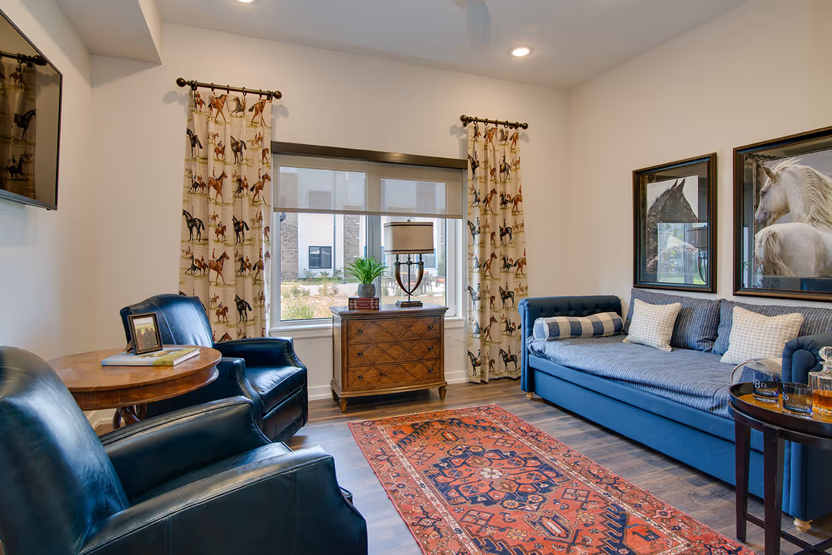 A cozy living room with a blue sofa adorned with pillows, two dark blue leather armchairs, a wooden side table with books and a framed photo, a wooden chest with a lamp and plant in front of a window with horse-patterned curtains, and two framed horse pictures on the wall. A red patterned rug covers part of the wooden floor.