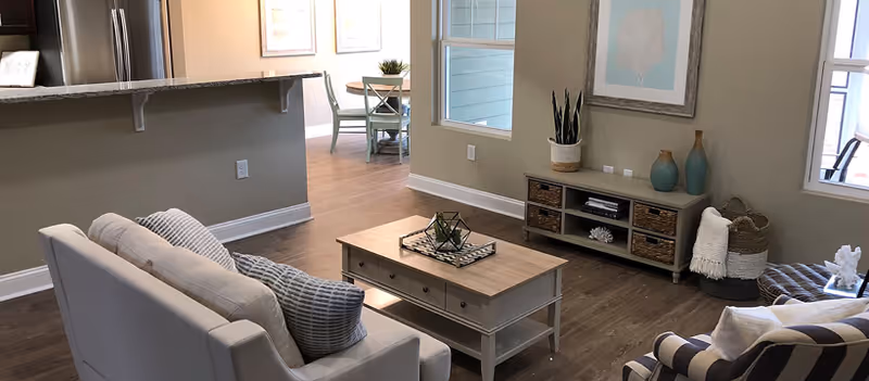 A cozy living room with a light gray sofa adorned with pillows, a wooden coffee table with decorative items, a striped armchair, and a TV stand with vases and plants. The room has wooden flooring and neutral-colored walls, with a view into a dining area and kitchen in the background.