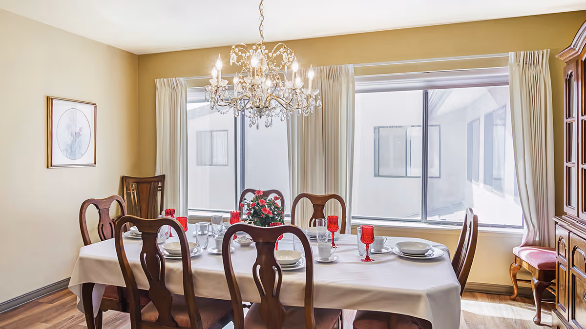 A dining room with a rectangular table covered with a white tablecloth, set with white plates, bowls, cups, and red goblets. There are six wooden chairs around the table. A chandelier with lit bulbs hangs above the table. The room has large windows with cream-colored curtains, wooden flooring, a framed floral picture on the wall, and a wooden cabinet with glass doors on the right side.