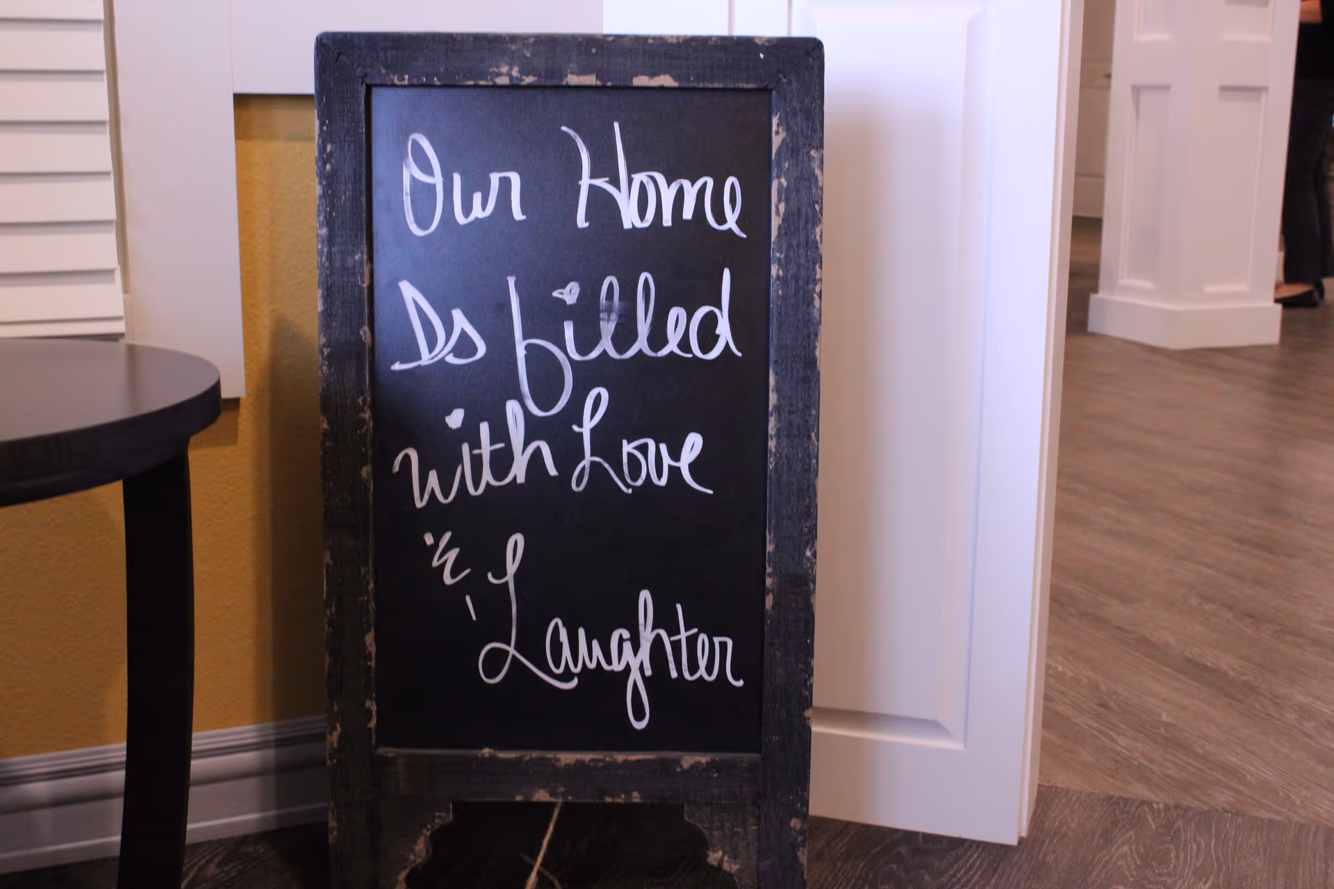 A small black chalkboard with a worn wooden frame standing on a wooden floor next to a round dark table. The chalkboard has white handwritten text that reads 'Our Home is filled with Love & Laughter'. The background shows part of a white door and a hallway with wooden flooring.