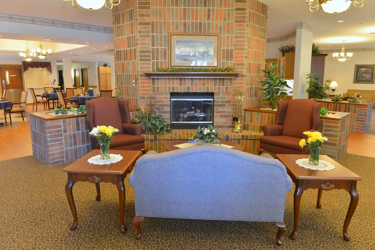 A cozy assisted living facility common area with a brick fireplace as the focal point. In front of the fireplace are two brown armchairs and a blue upholstered loveseat, arranged around a wooden coffee table with a floral centerpiece. Two wooden side tables with vases of yellow and white flowers flank the loveseat. The room has carpeted flooring near the seating area and wood flooring in the dining area visible in the background, which contains several tables and chairs. The space is warmly lit with chandeliers and decorated with plants and framed artwork.