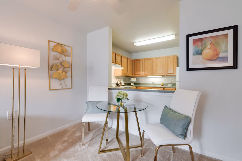 A small dining area with a round glass table and two white chairs with blue-green cushions. A floor lamp with a white shade stands to the left, and two pieces of wall art are visible: one with gold leaves and another framed painting of a pear. In the background, a kitchen with wooden cabinets and a countertop is visible.