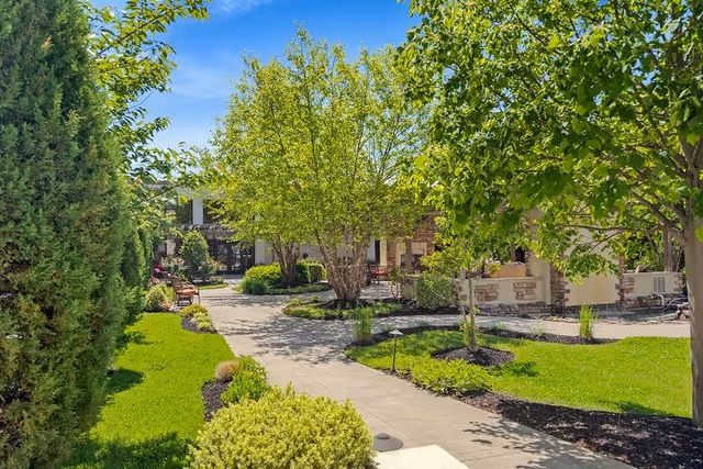 A sunny outdoor garden area with a paved walkway winding through green grass, trees, and shrubs. Benches are placed along the path, and a building with stone and beige walls is visible in the background under a clear blue sky.