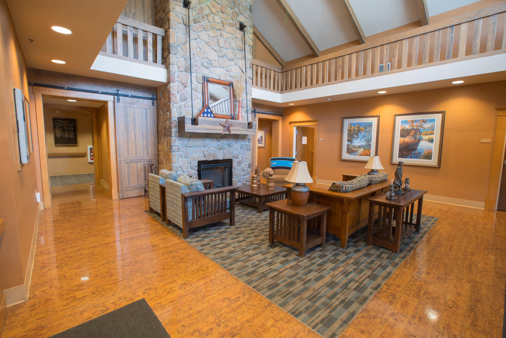 A cozy living room area in a senior living facility with a high vaulted ceiling and a large stone fireplace. The room features wooden furniture including sofas, armchairs, side tables with lamps, and a coffee table on a patterned rug. The walls are painted warm beige and decorated with framed landscape paintings. There is a wooden sliding barn door and a balcony railing above the fireplace.