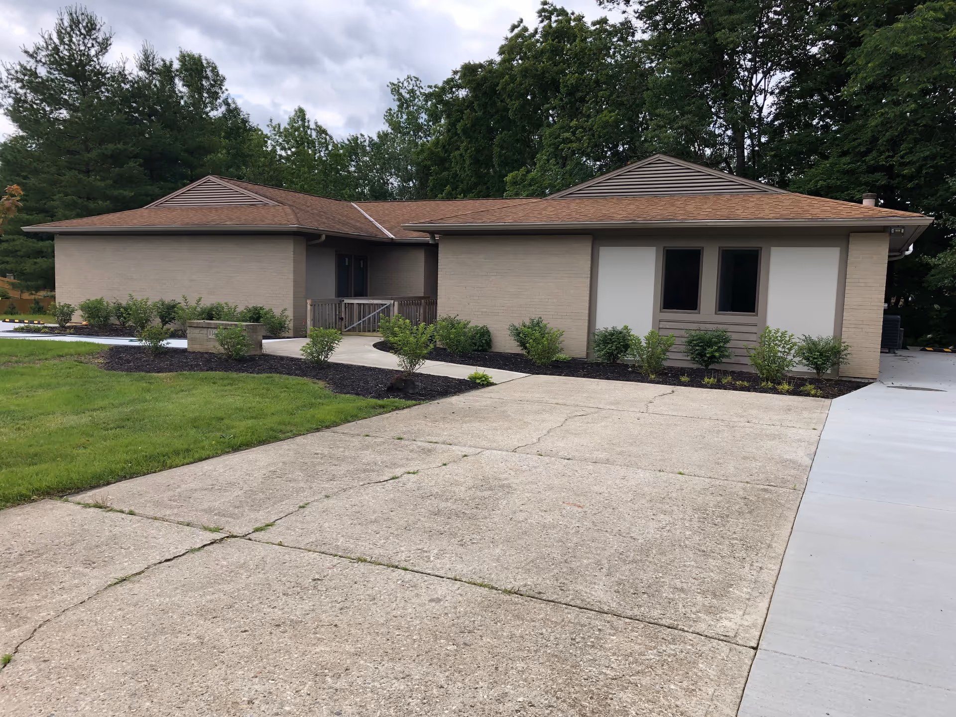 Single-story beige brick building with a brown roof surrounded by green trees and shrubs, with a concrete driveway and walkway leading to the entrance.