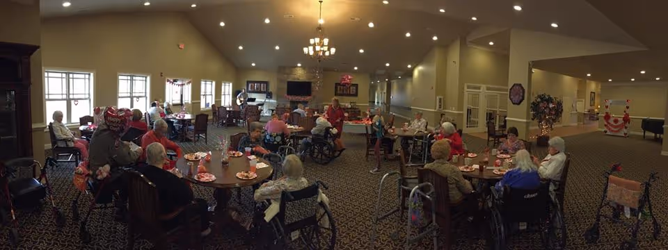 A large common area in an assisted living facility with multiple round tables where elderly residents are seated, some in wheelchairs and others in regular chairs. The room is decorated with Valentine’s Day themed items, including heart-shaped balloons and red decorations. There is a chandelier hanging from the ceiling, a fireplace on the left side, and a television mounted on the far wall. The carpet has a patterned design, and the room has large windows letting in natural light.