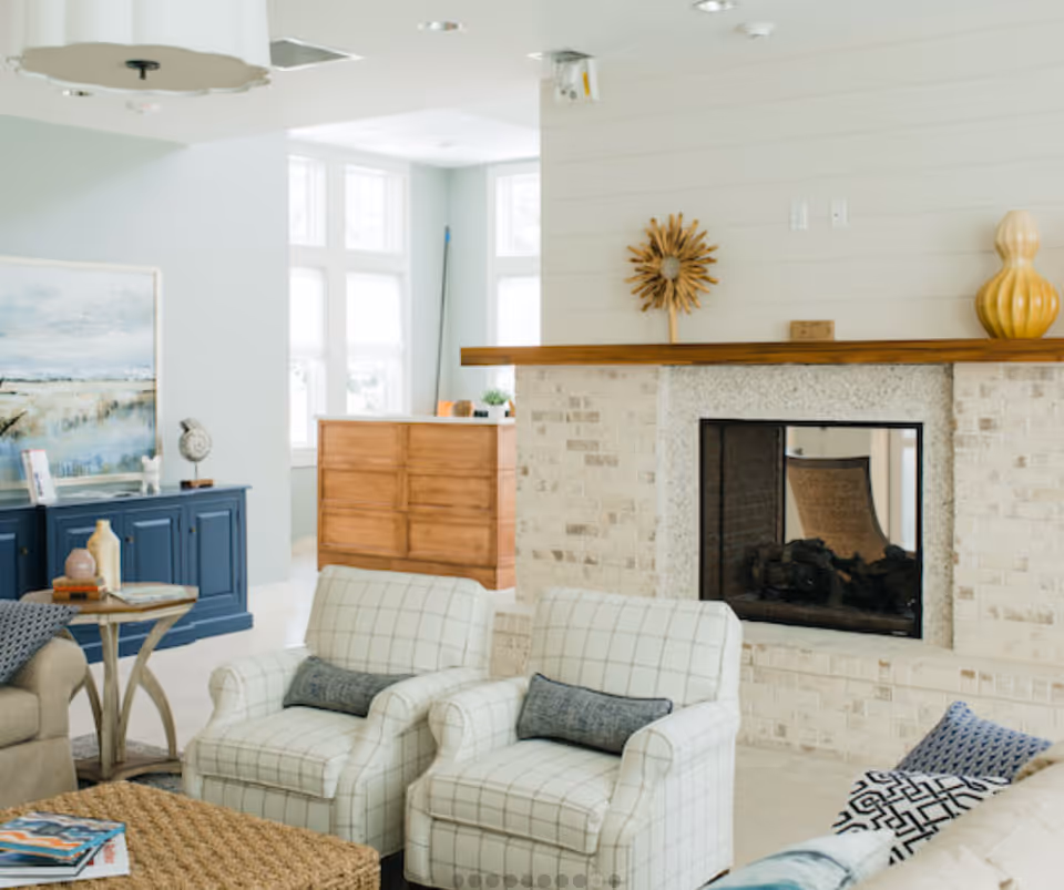 A cozy living room with two white checkered armchairs, a beige sofa with patterned pillows, a woven coffee table, and a brick fireplace with a wooden mantel decorated with a sunburst ornament and a yellow vase. In the background, there is a blue sideboard with decorative items and a large window letting in natural light.