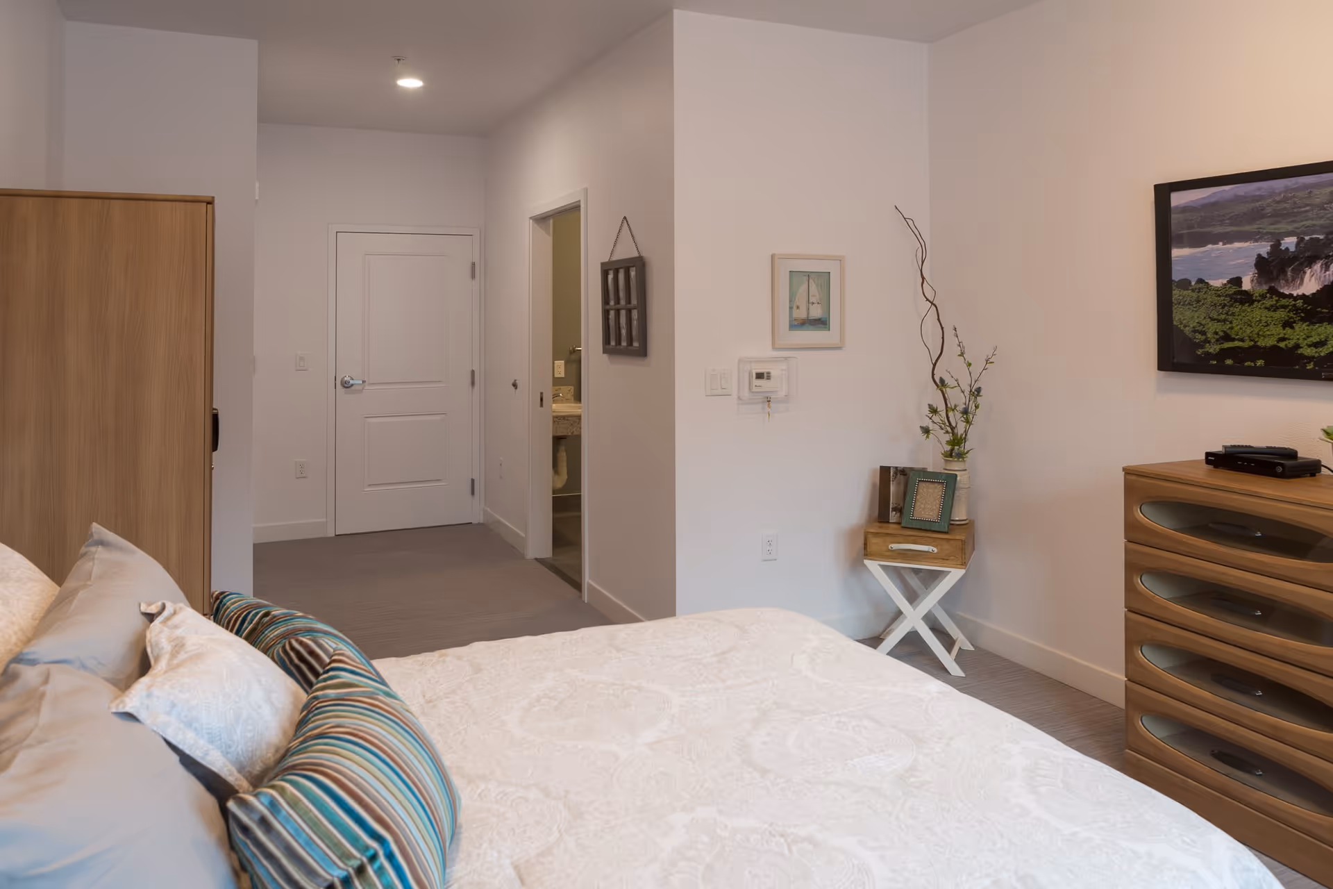 A bedroom in a senior living facility with a neatly made bed featuring multiple pillows, a wooden dresser with a TV on top, a small side table with decorative items and a framed picture on the wall. There is a doorway leading to a bathroom and another door leading out of the room.