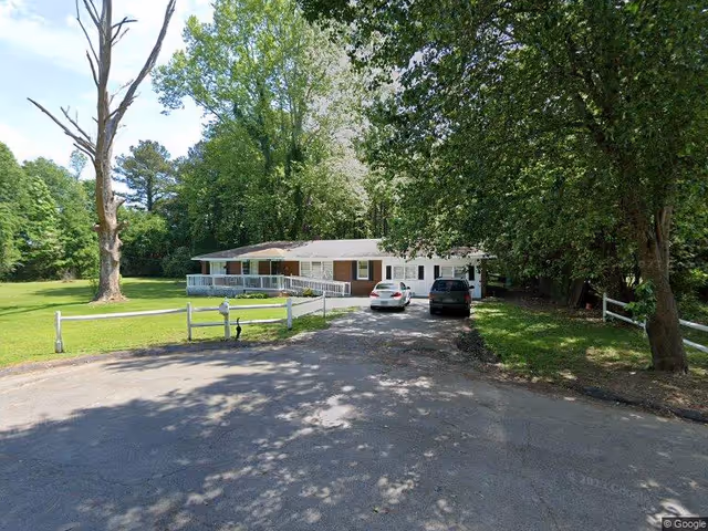 Single-story building with a white railing and two cars parked in front, surrounded by large green trees and a grassy lawn under a clear sky.