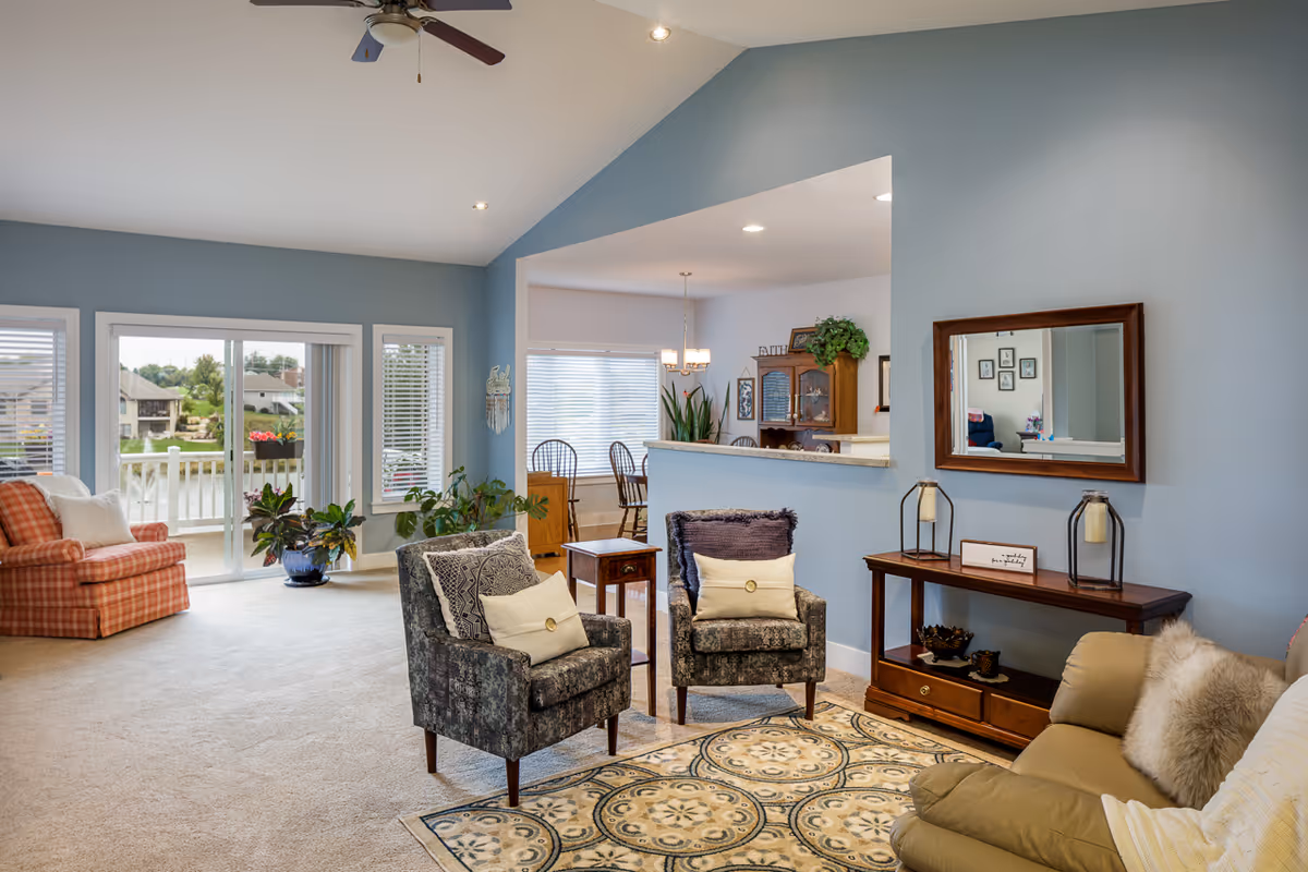 A cozy living room with light blue walls, beige carpet, and a ceiling fan. The room features two patterned armchairs with cushions, a beige sofa with pillows, a wooden console table with decorative items and a mirror above it. There is a patterned area rug on the floor. In the background, a dining area with a wooden table and chairs is visible through a large open wall. Large windows and a sliding glass door provide natural light and a view of an outdoor patio and neighboring houses.