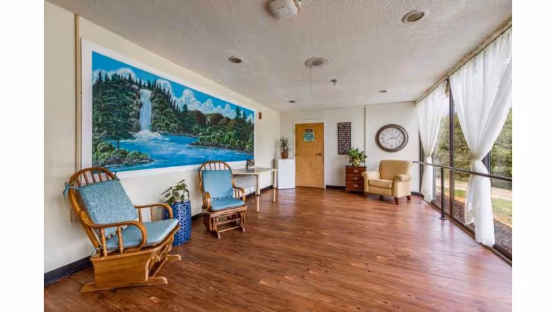 Sunlit common sitting area with wooden rocking chairs, a large nature mural, floor-to-ceiling windows with sheer curtains, and wood flooring.