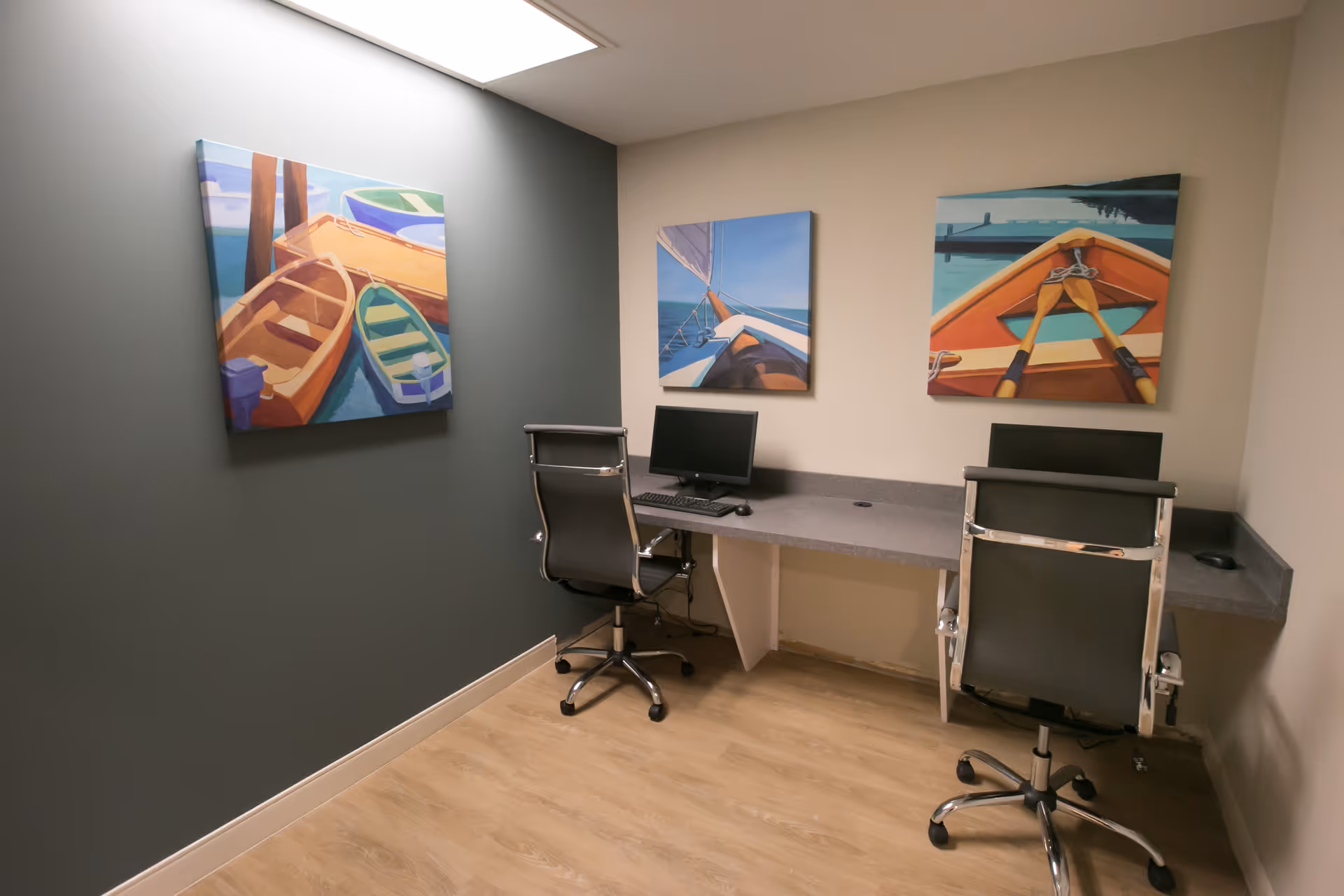 Small computer workstation with two office chairs and colorful boat paintings on the walls.