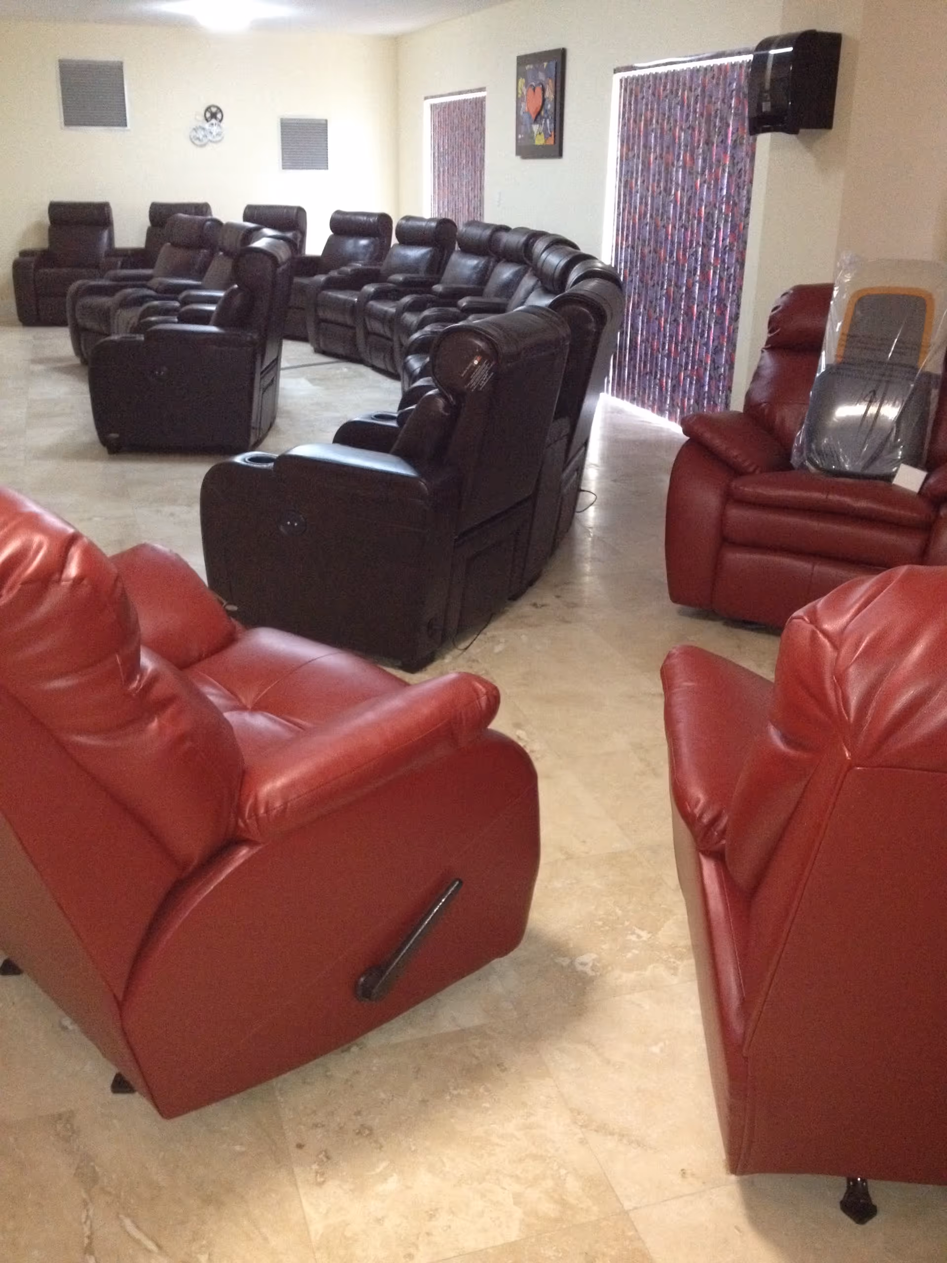 A room with multiple recliner chairs arranged in rows facing the same direction. The chairs are mostly dark brown with a few red ones in the foreground. The room has light-colored tiled flooring, two windows with patterned curtains, and a wall-mounted dispenser. There is a framed artwork on the wall between the windows.