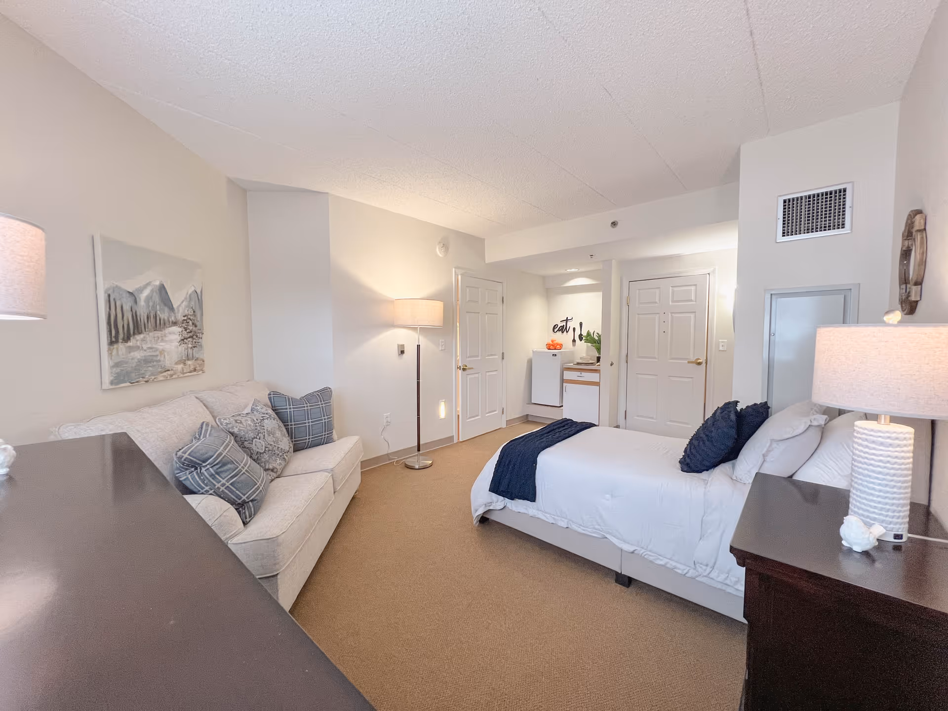 A cozy senior living studio apartment featuring a bed with white linens and dark blue pillows, a beige sofa with patterned cushions, a floor lamp, a bedside table with a lamp, and a small kitchenette area in the background with a mini fridge and a sink. The walls are light-colored, and there is a landscape painting above the sofa.